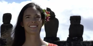 Primer plano de una mujer sonriente con flores en el cabello, frente a las estatuas Moái en la Isla de Pascua.