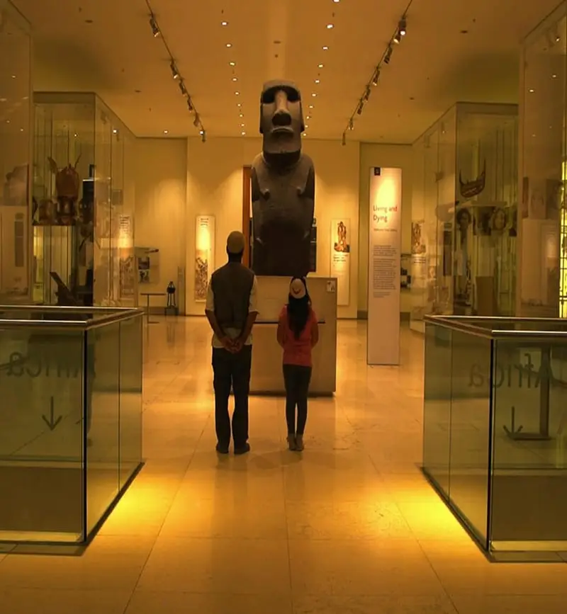 Dos personas de pie en un museo, observando un moái, una estatua de piedra de la Isla de Pascua.